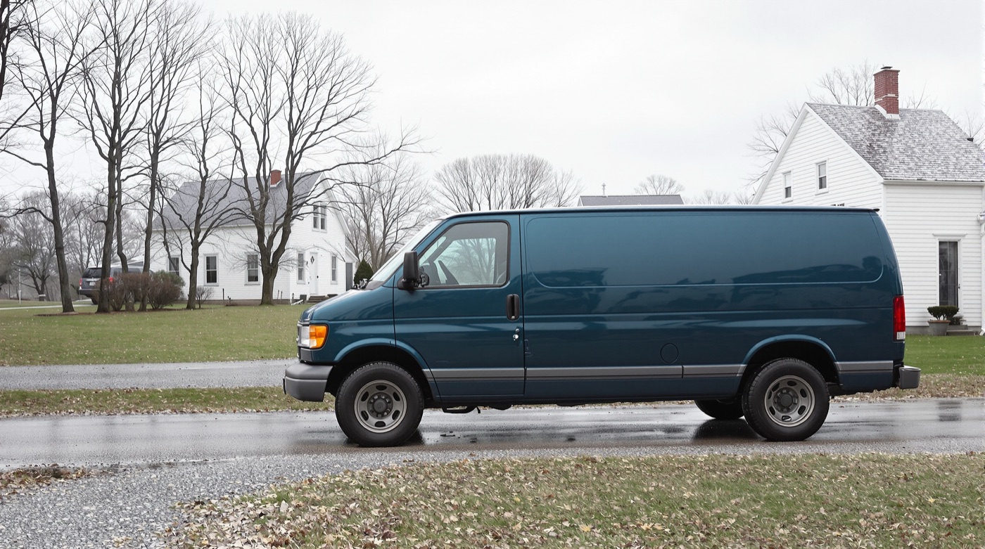 A plain deep teal work van parked on a wet gravel drive beside a white farmhouse, bare trees and a grey sky behind.