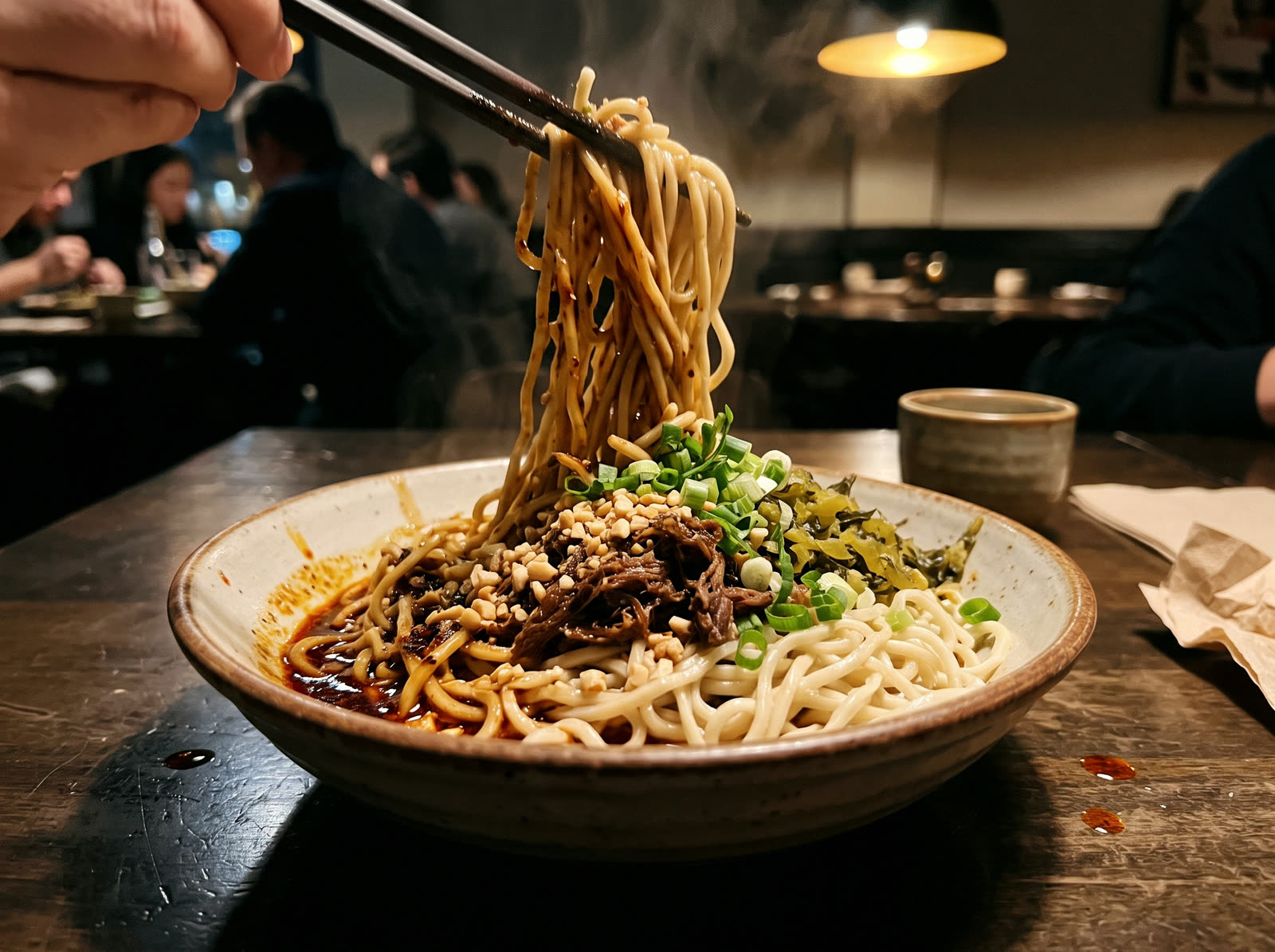 Chopsticks lifting noodles from a bowl of dan dan noodles with chili oil, beef and peanuts