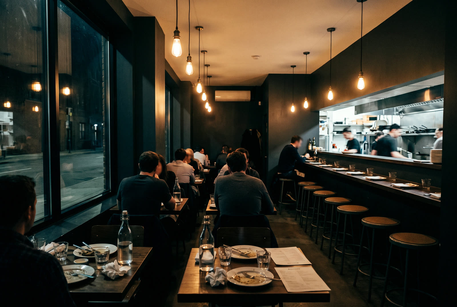 A narrow dining room at night with pendant lights and a row of stools facing an open kitchen