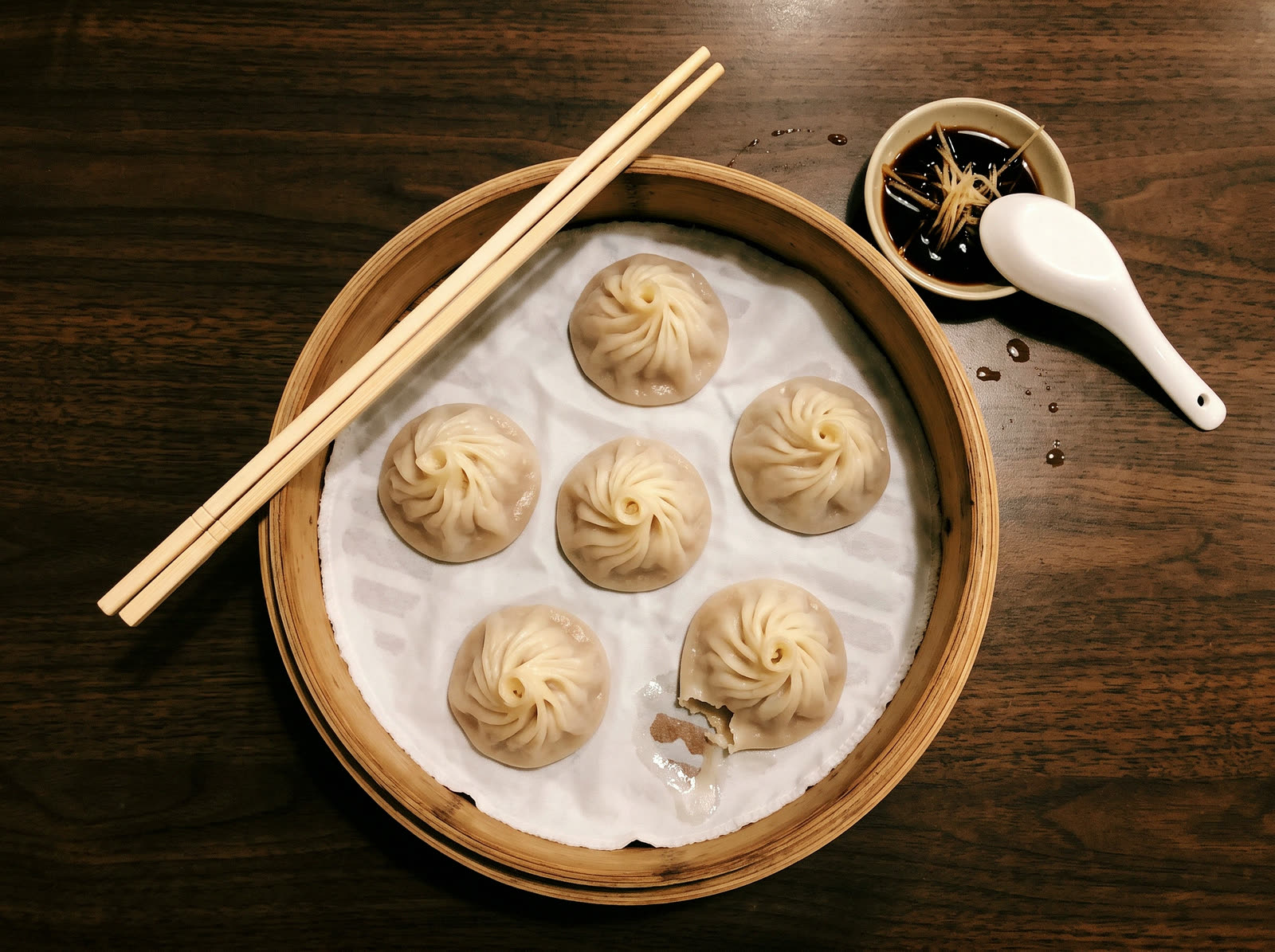 Six soup dumplings in a bamboo steamer, seen from above, with black vinegar, ginger and a spoon