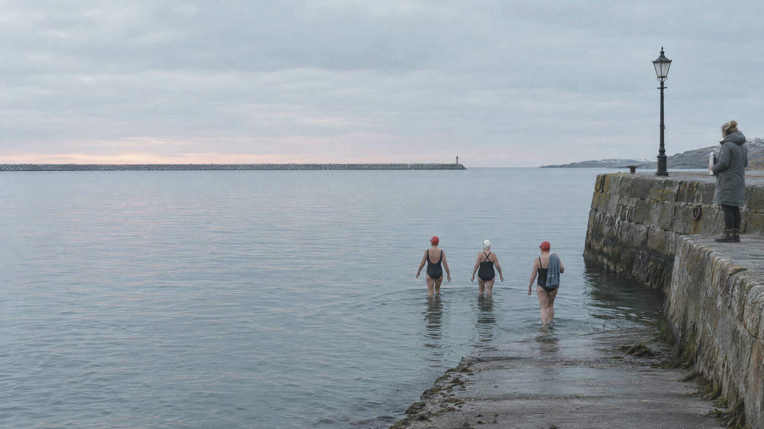 Three swimmers in red and white caps wading into the harbour off the slipway at dawn, a member with a flask watching from the quay