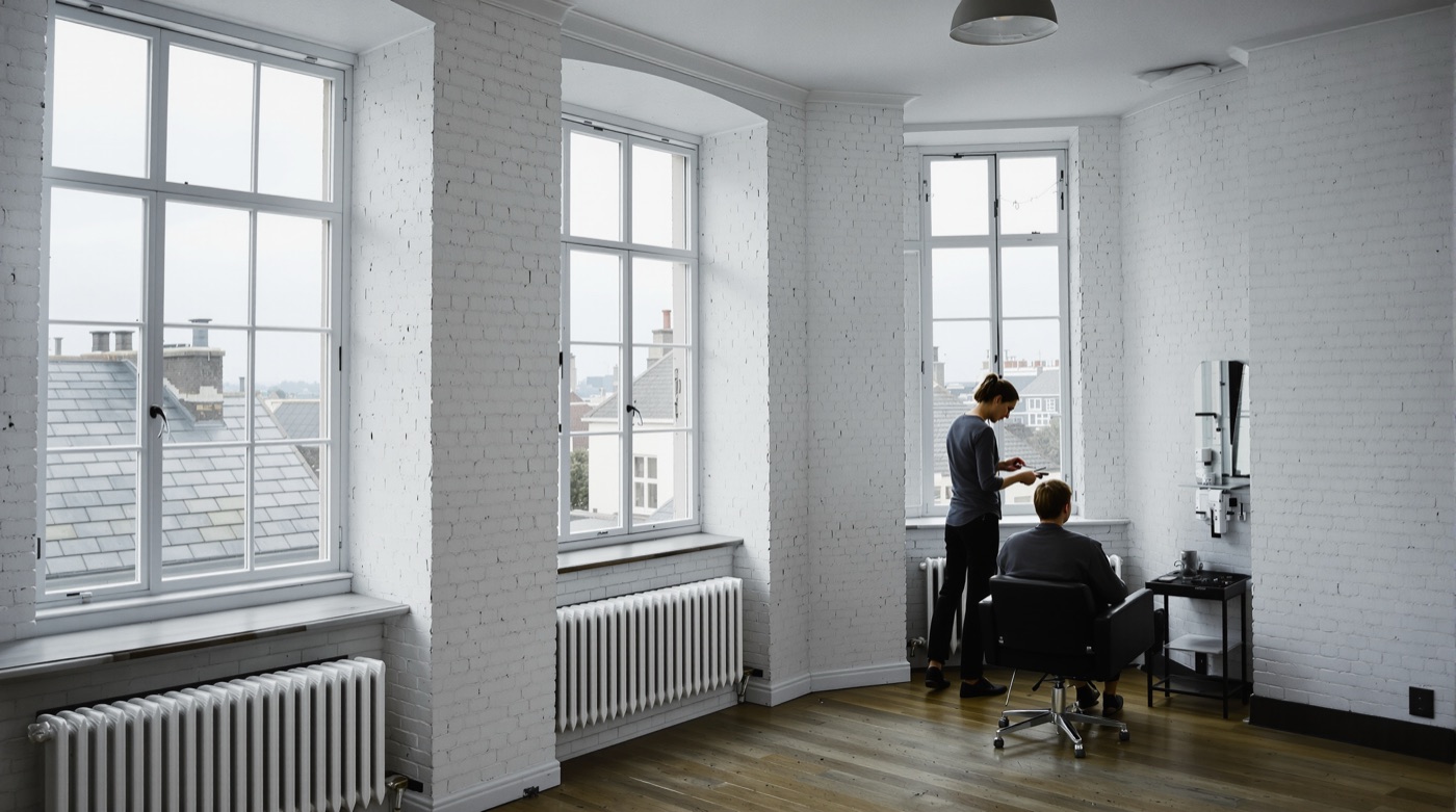 A wide white brick room with tall windows over slate rooftops; far off by the right window a hairdresser cuts a seated client's hair