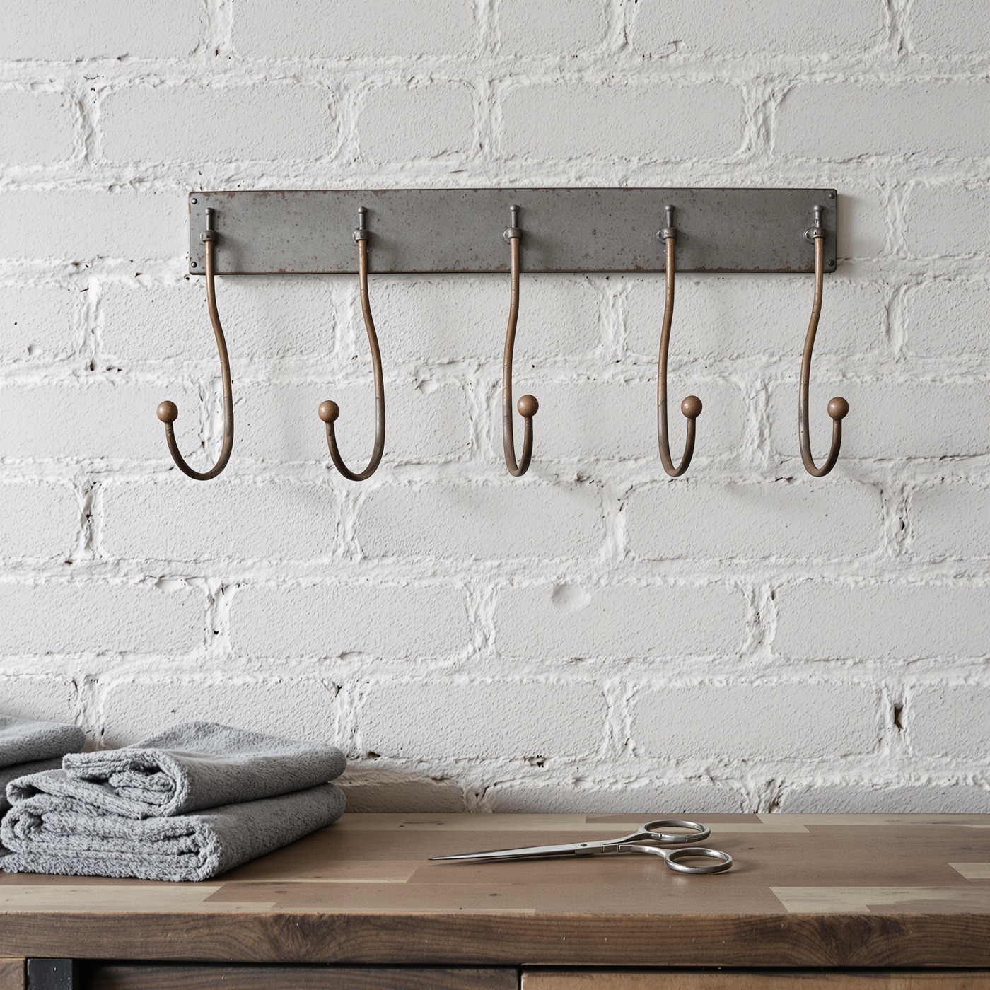 Old iron hooks on a whitewashed brick wall above a wooden counter with folded grey towels and a pair of scissors