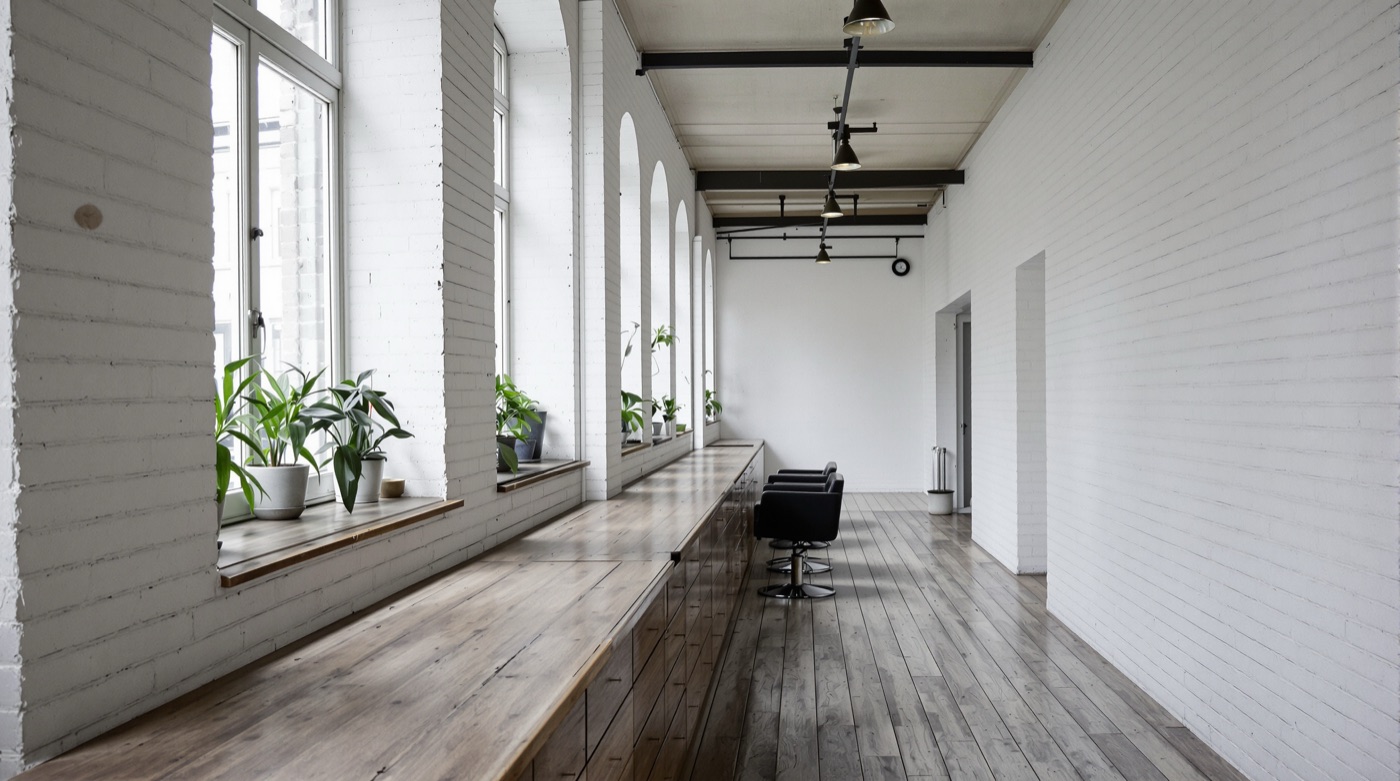 A long worn wooden counter with drawers beside tall windows with plants on the sills, two empty salon chairs at the far end of a white brick room