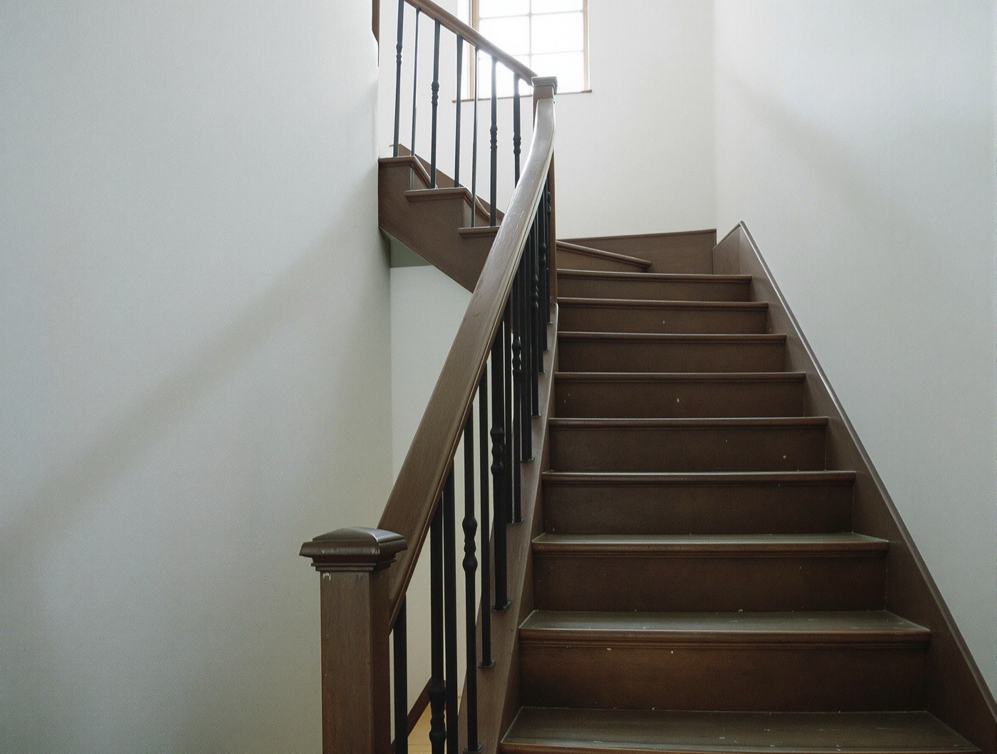 A narrow wooden staircase with a dark handrail rising to a window on the floor above